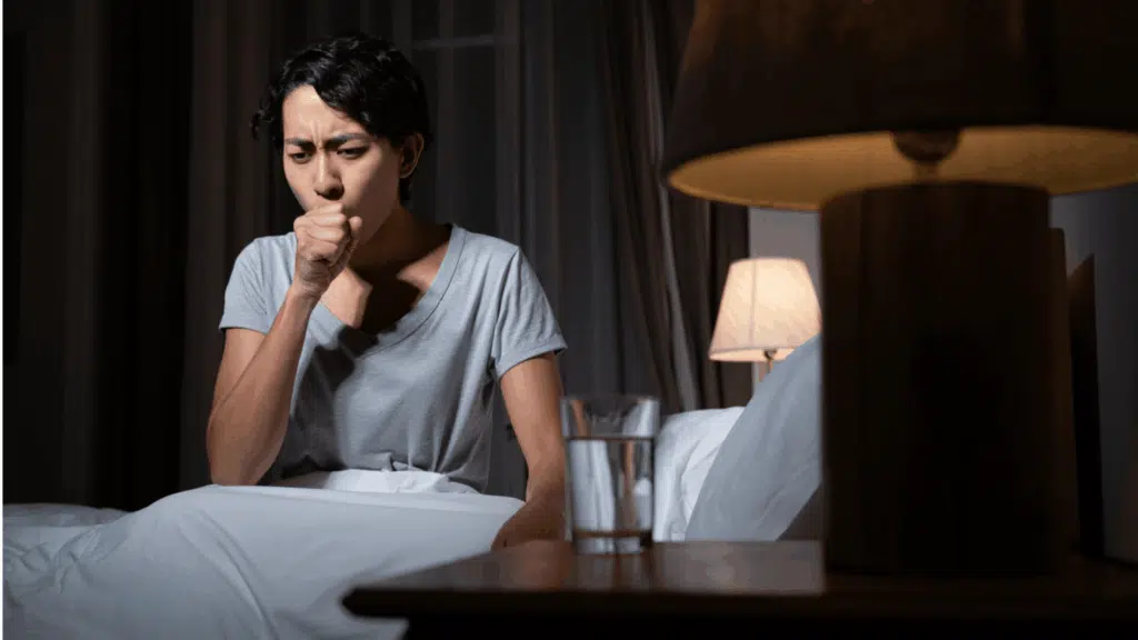 person sitting up in bed at night coughing and distressed, struggling to breathe in a dimly lit bedroom (1)