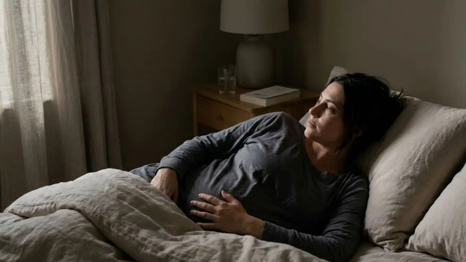 a woman in a sleep shirt lies awake in a dimly lit bedroom, resting her hand on her early pregnancy bump