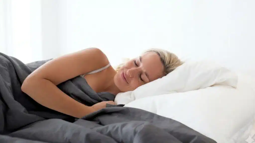 freckled woman with blonde hair sleeps soundly in a bed, covered by a grey duvet and white pillows