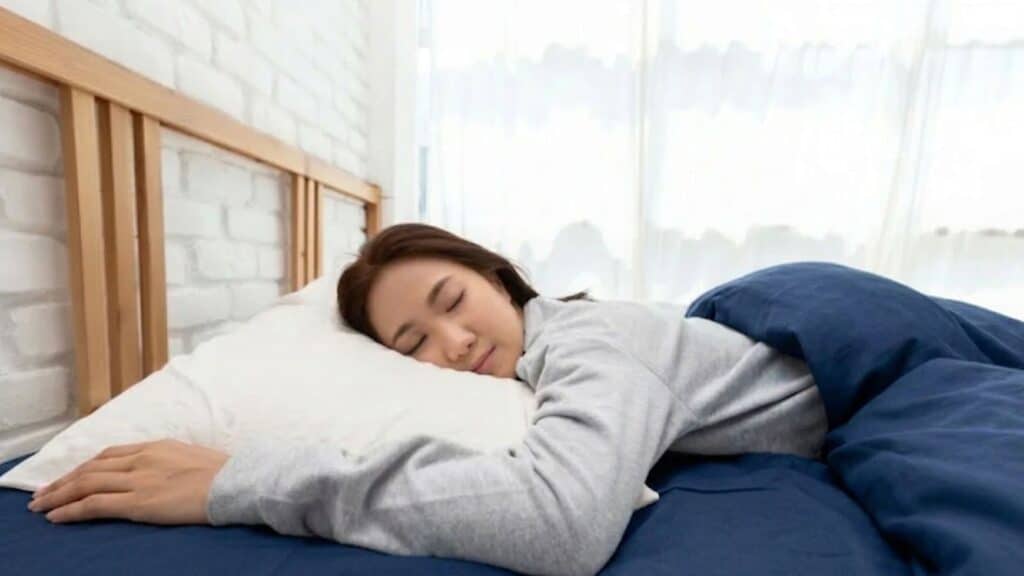 woman with dark hair sleeps peacefully on a white pillow, covered by a grey blanket and matching bedsheets