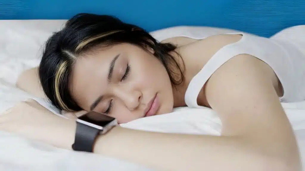 young woman with dark hair and blonde highlights sleeps on white bedding while wearing a black smartwatch for tracking