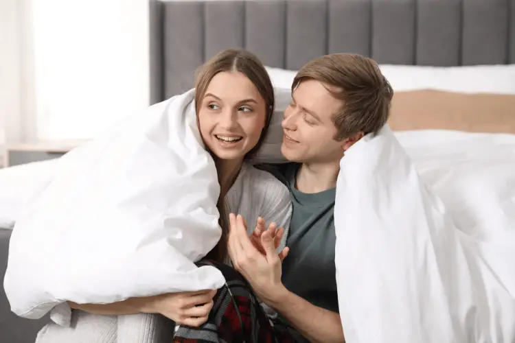 Couple sitting on bed wrapped in white duvet in cozy bedroom setting