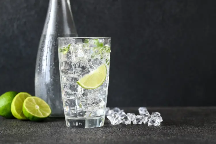 Glass of sparkling water with lime wedge and ice on dark textured surface