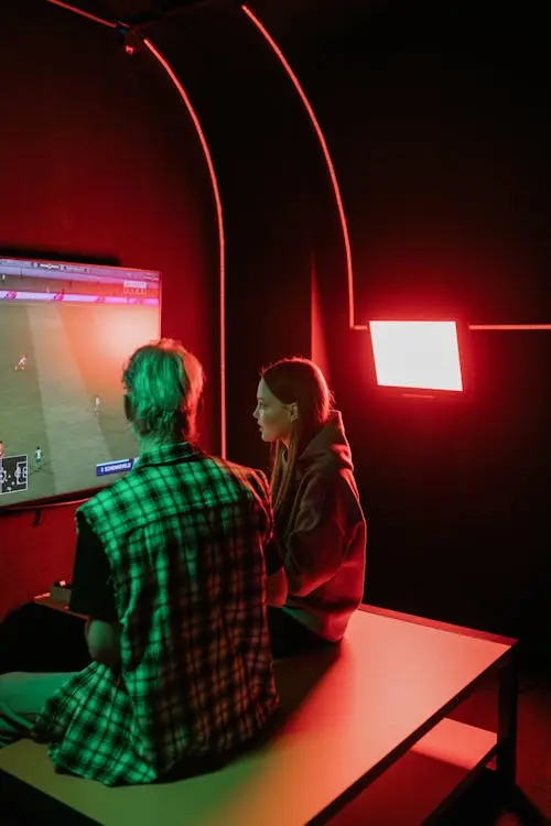 Two people playing video game on large screen in dark room with red lighting