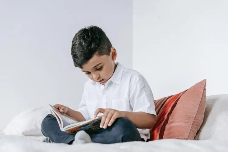Young boy in white shirt reading book on bed with striped pillows