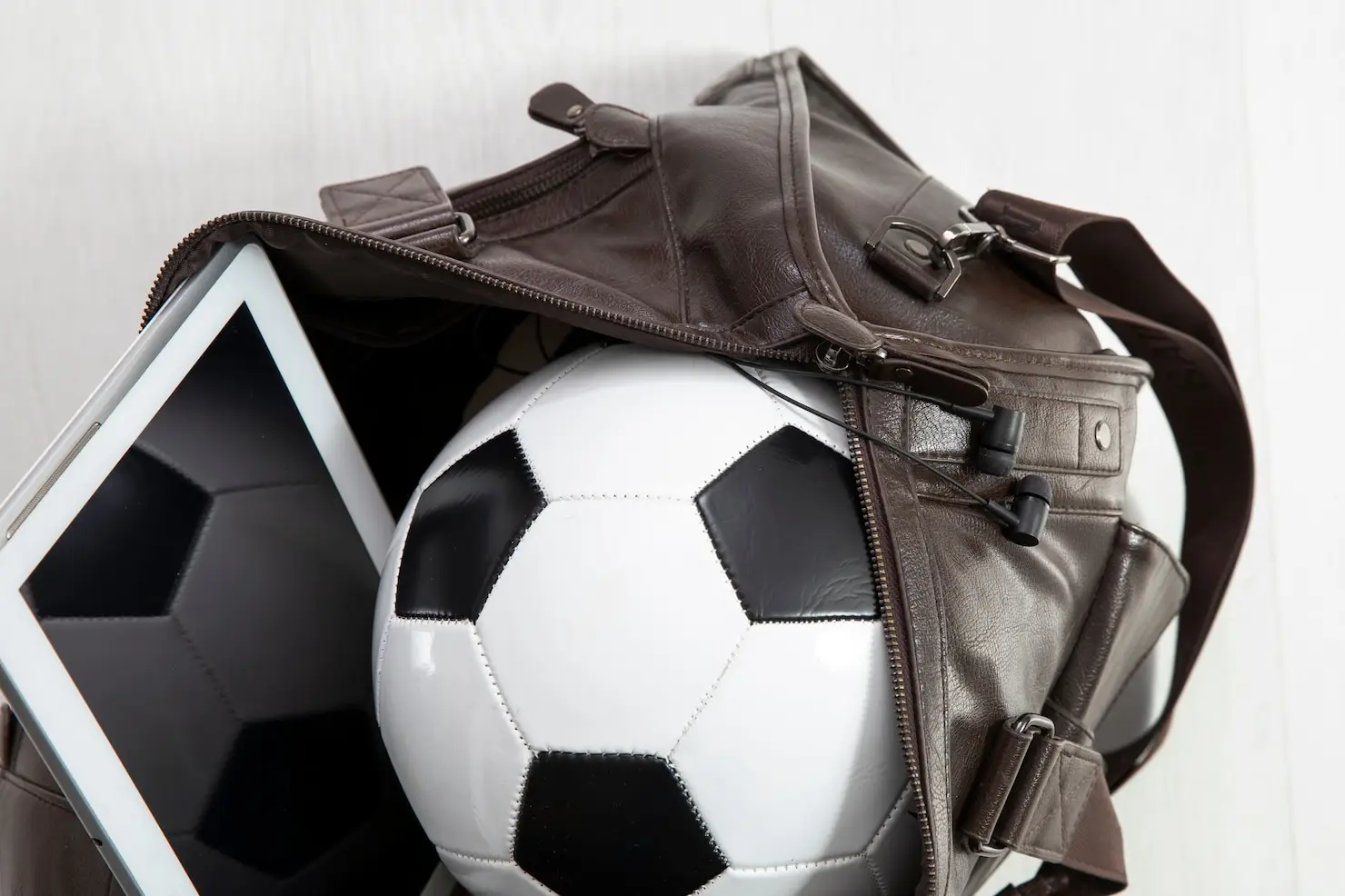 Soccer ball and tablet inside open leather duffel bag on light floor