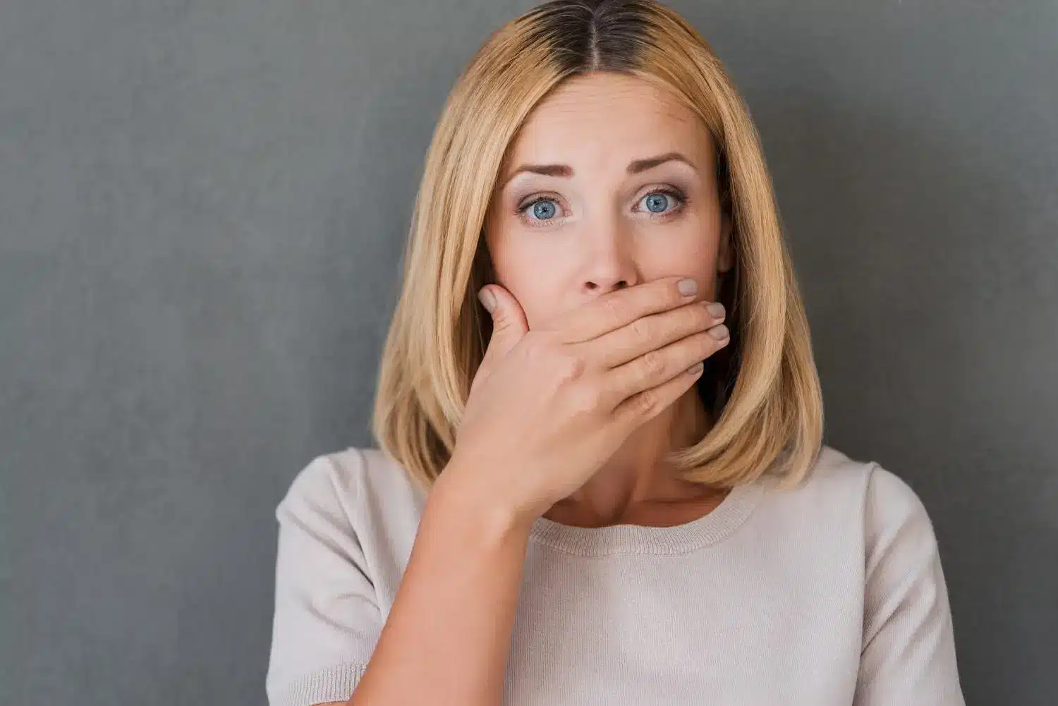 Woman with blonde hair covering mouth with hand against gray background