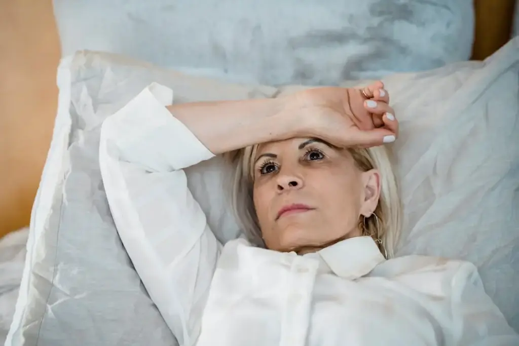 Woman lying in bed with hand on forehead wearing a white blouse in a bright room