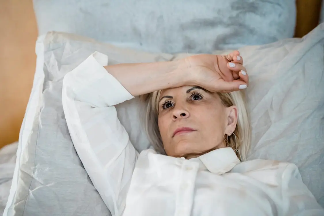 Woman lying in bed with hand on forehead wearing a white blouse in a bright room