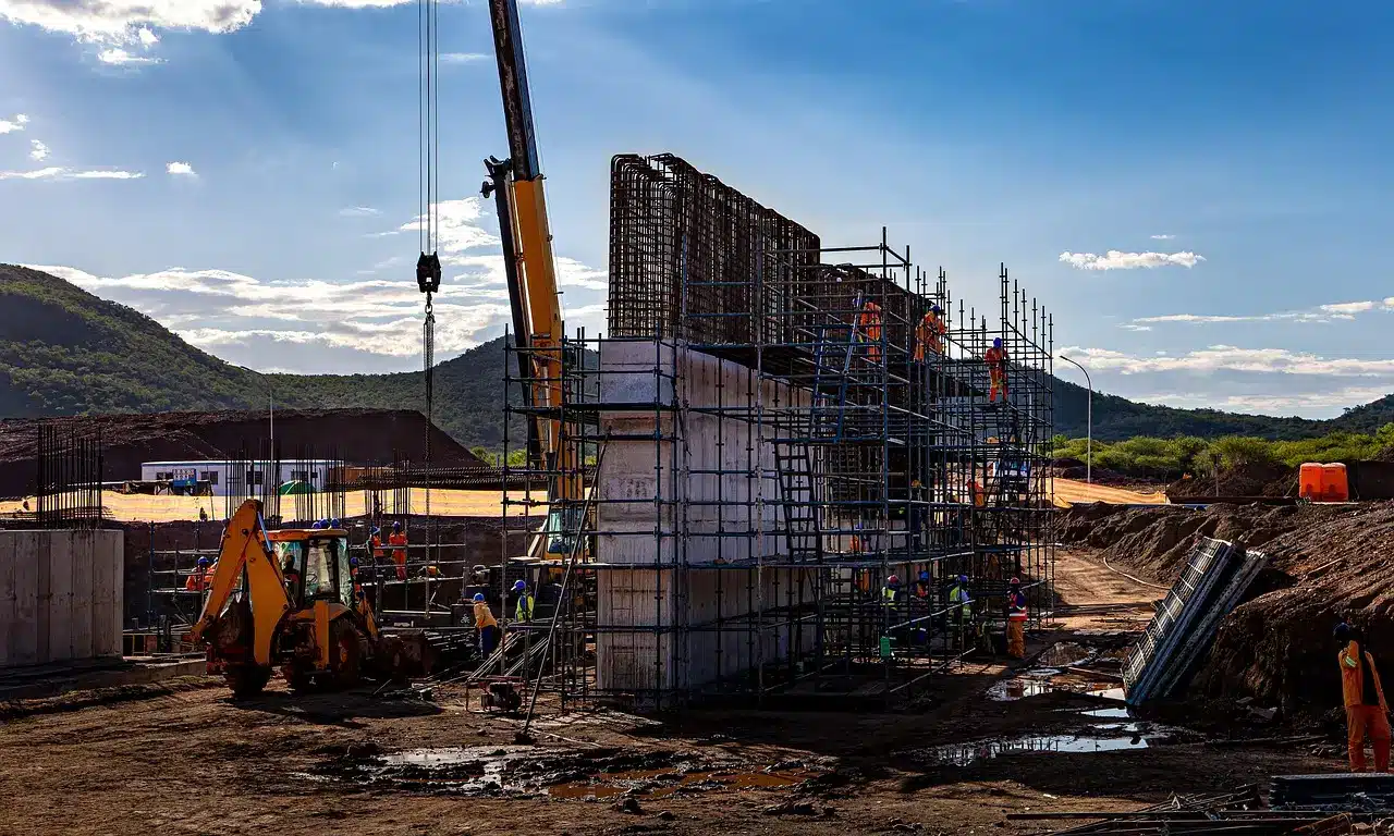 Construction site with scaffolding and workers near crane, set against hills and clear blue sky