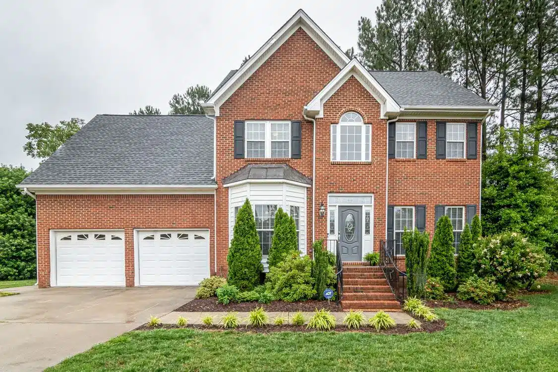 Red brick two-story house with black shutters and manicured shrubs in a suburban setting