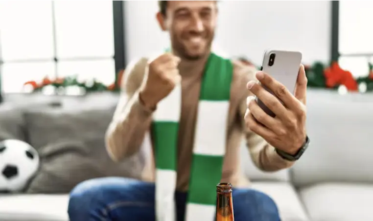 Man celebrating while looking at smartphone, wearing green and white scarf in living room with football decor