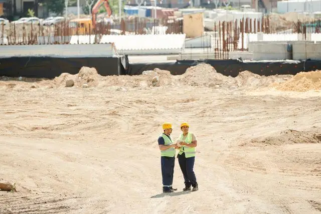 Two construction workers in safety gear discussing plans on a large construction site