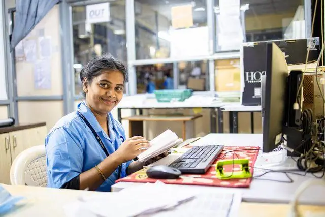 Smiling woman in blue uniform working at cluttered office desk with computer and papers