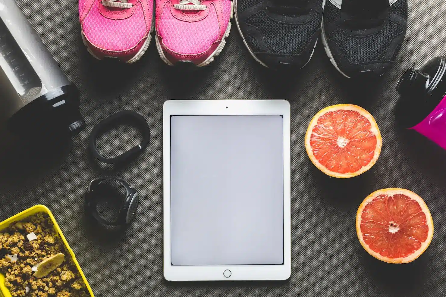 Tablet with pink and black sneakers, grapefruit halves, fitness trackers, and protein shaker on gray surface