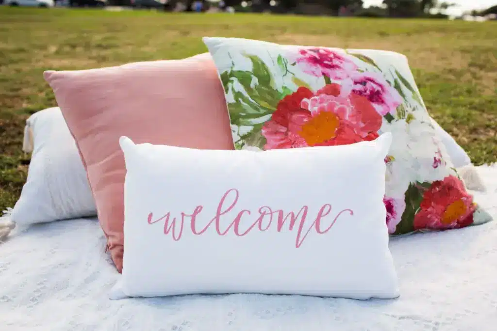 White welcome pillow with floral and peach cushions on lawn blanket in sunny outdoor setting