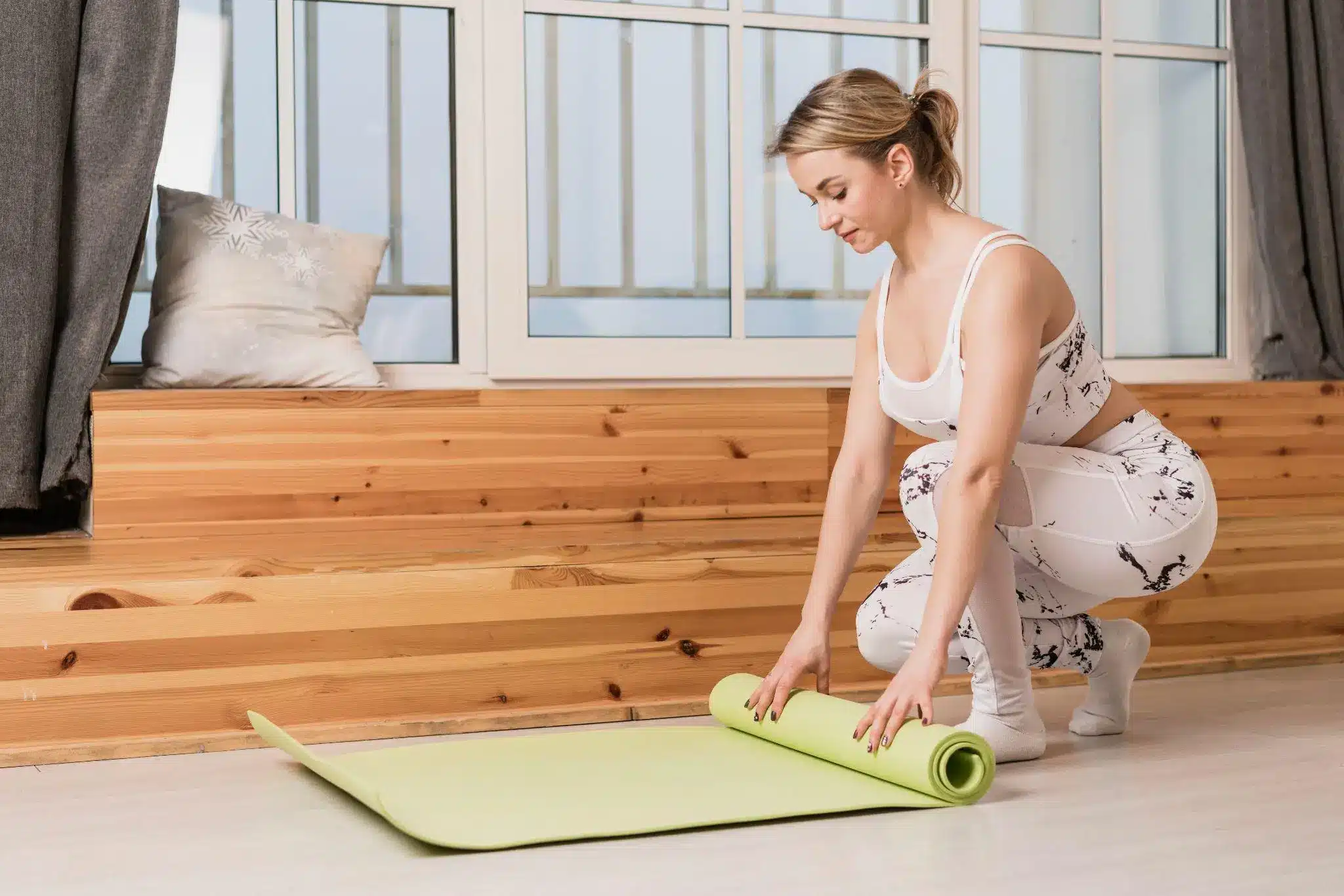 Woman in workout attire rolling a green yoga mat in a bright room