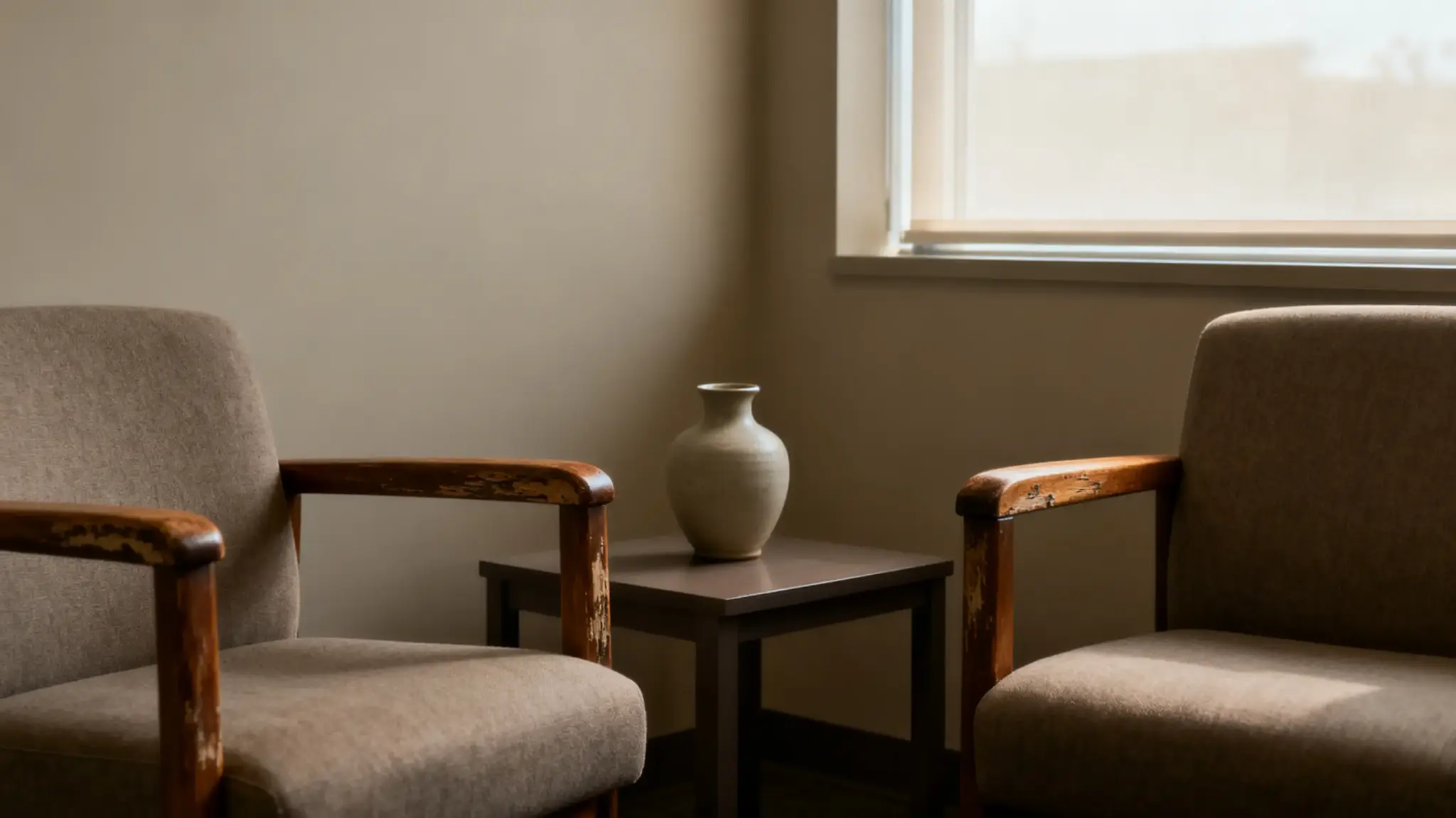 Two beige chairs with wooden arms flanking small table with ceramic vase in sunlit room