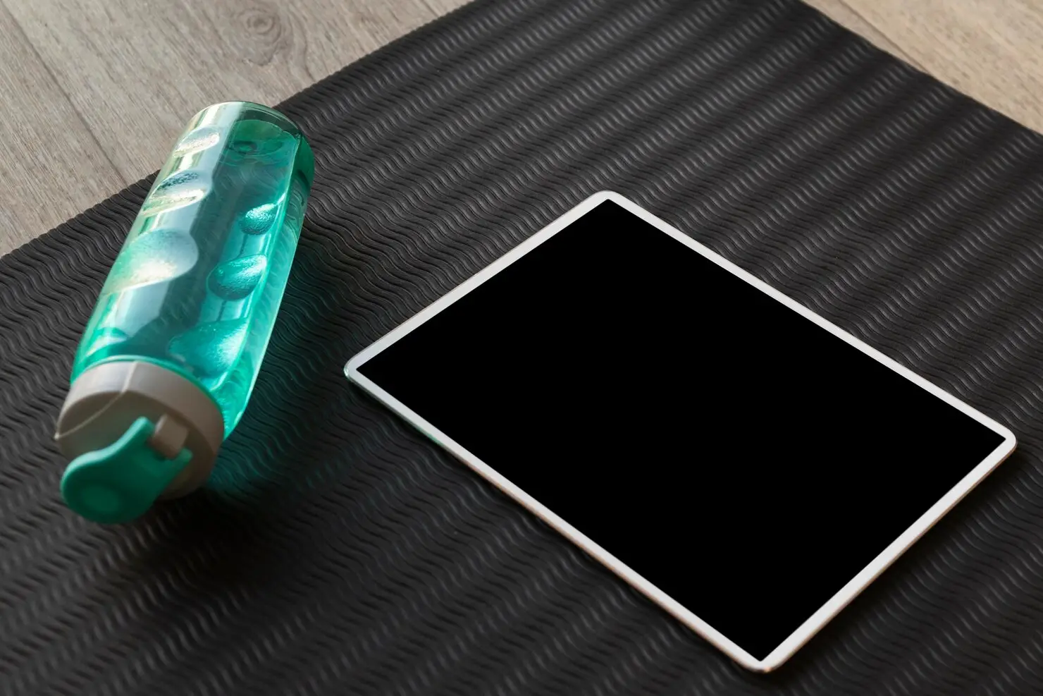 Tablet and turquoise water bottle on textured black fitness mat on wooden floor