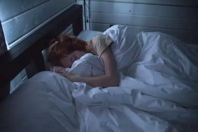 Person sleeping under white blanket in a dimly lit room with wooden walls