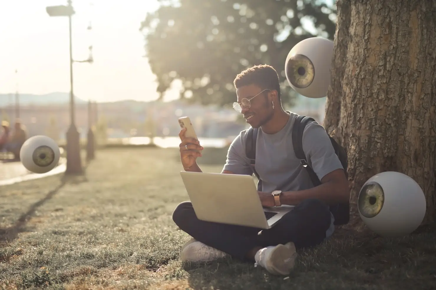Young man using laptop and phone under tree with large floating eyeballs in park setting