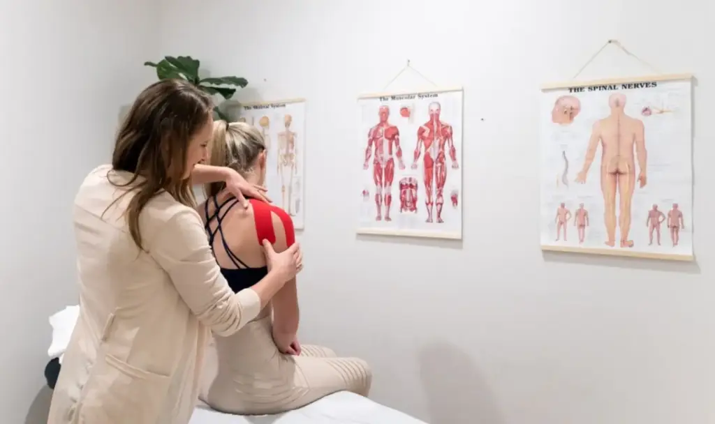 Chiropractor applying kinesiology tape on woman's shoulder in therapy room with anatomy charts