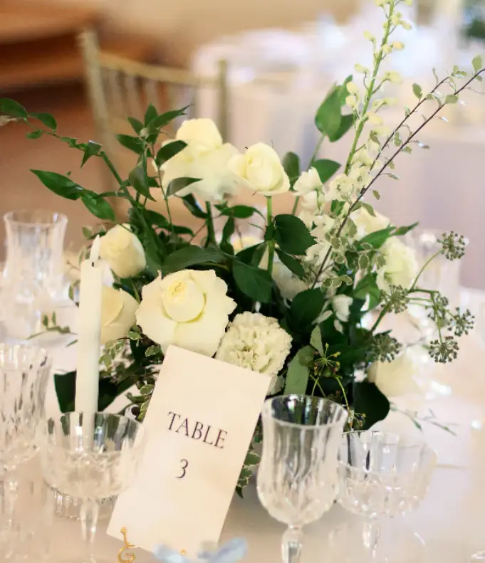 White roses and greenery centerpiece with table number card and crystal glasses on wedding reception table