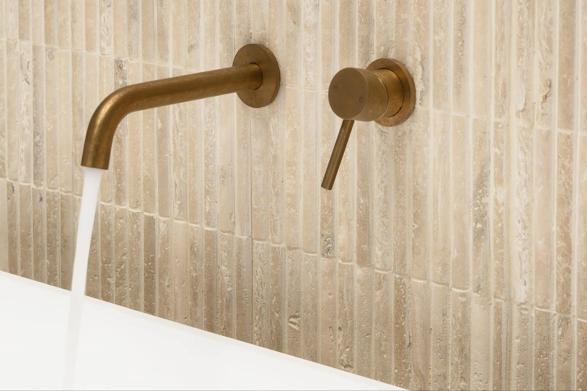 Bronze bathroom faucet with running water against textured beige tiled wall