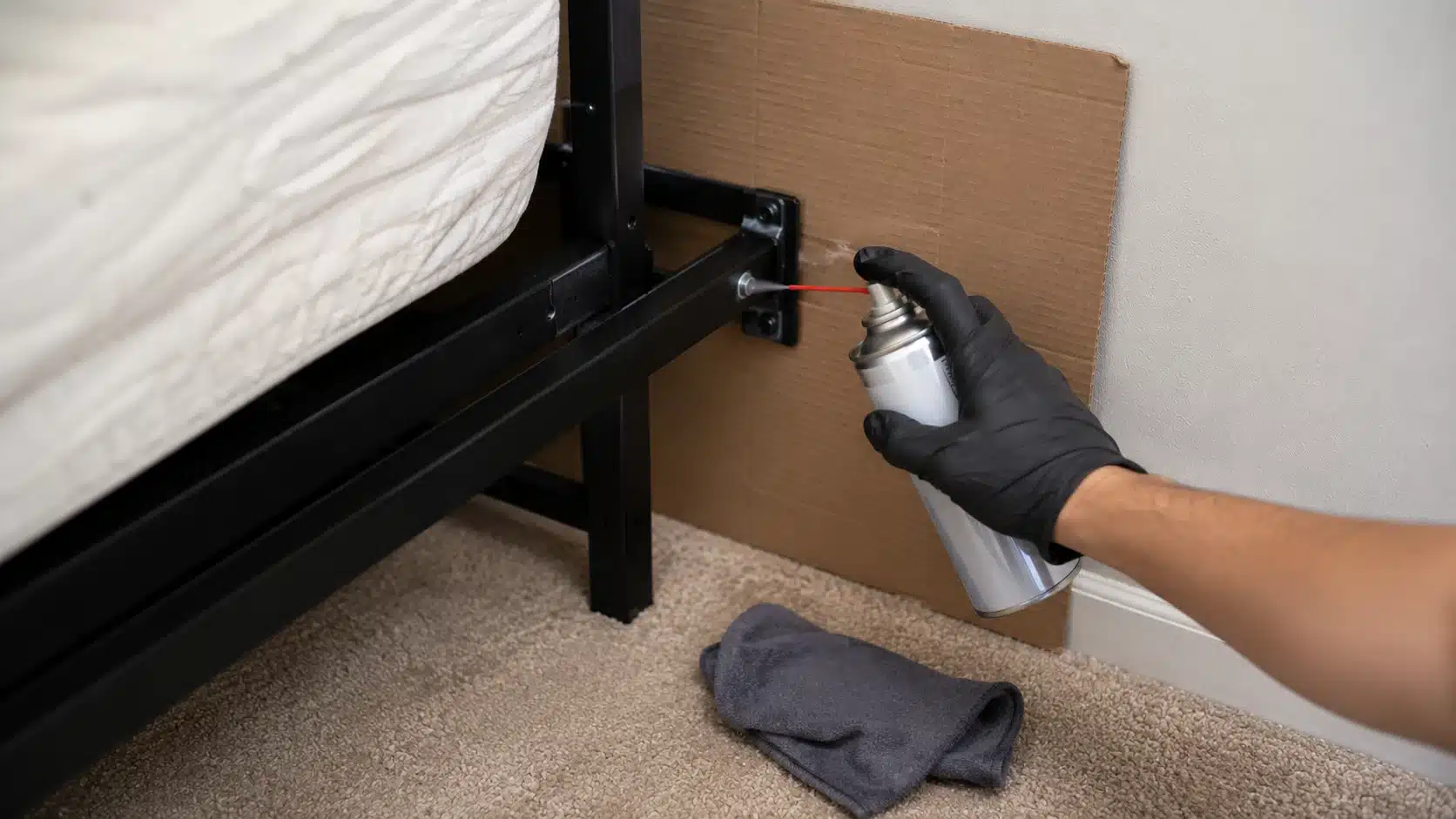 gloved hand applying silicone lubricant to a metal bed frame joint with cardboard behind it