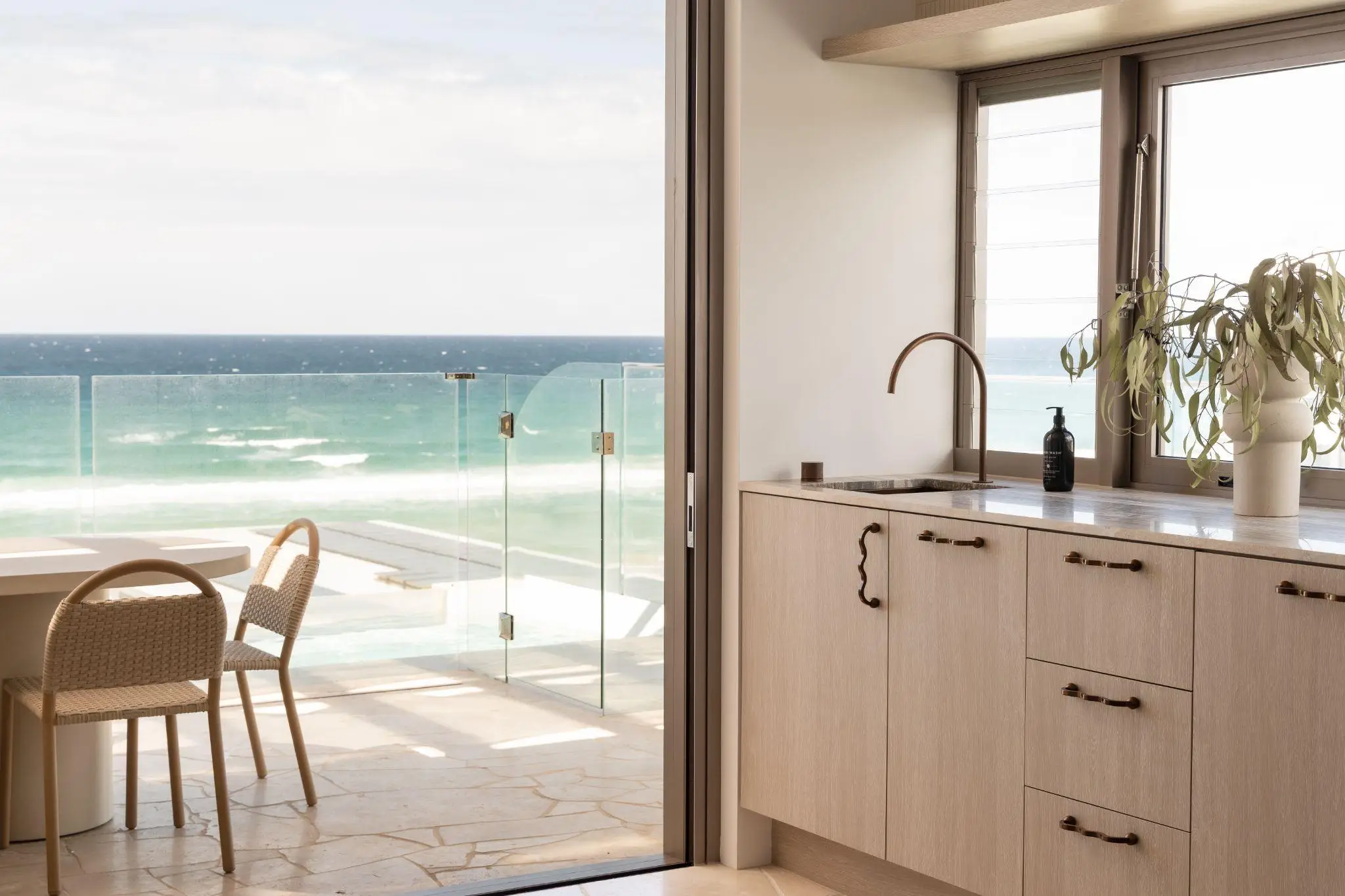 Modern kitchen with wooden cabinets, ocean view through glass doors, wicker chairs by round table