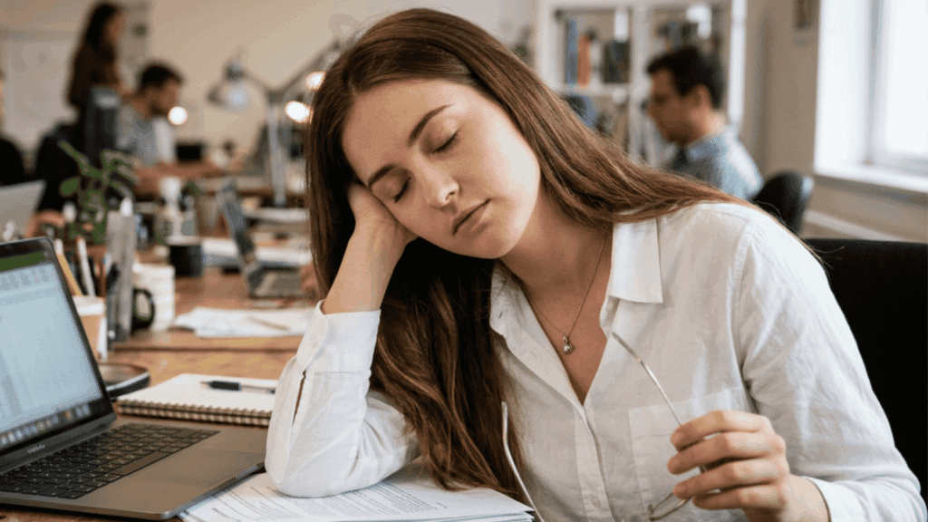 Sleep-deprived woman dozing at an office desk beside a laptop and papers