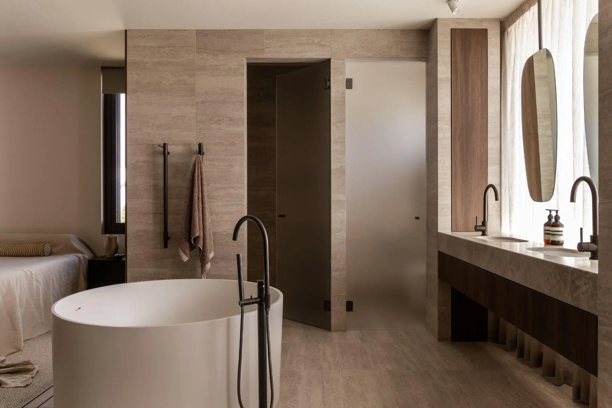 Modern minimalist bathroom featuring freestanding tub, dual vanities, and beige tiled walls in soft lighting
