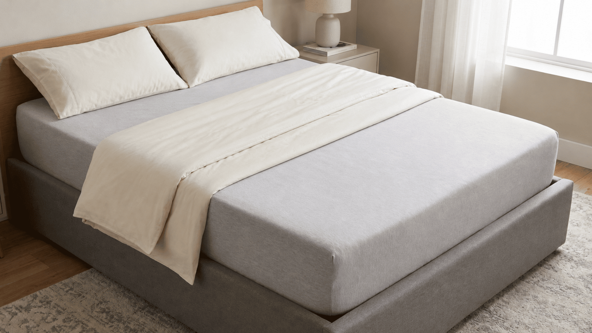 neatly made gray bed with cream bedding beside a bright window in a calm bedroom