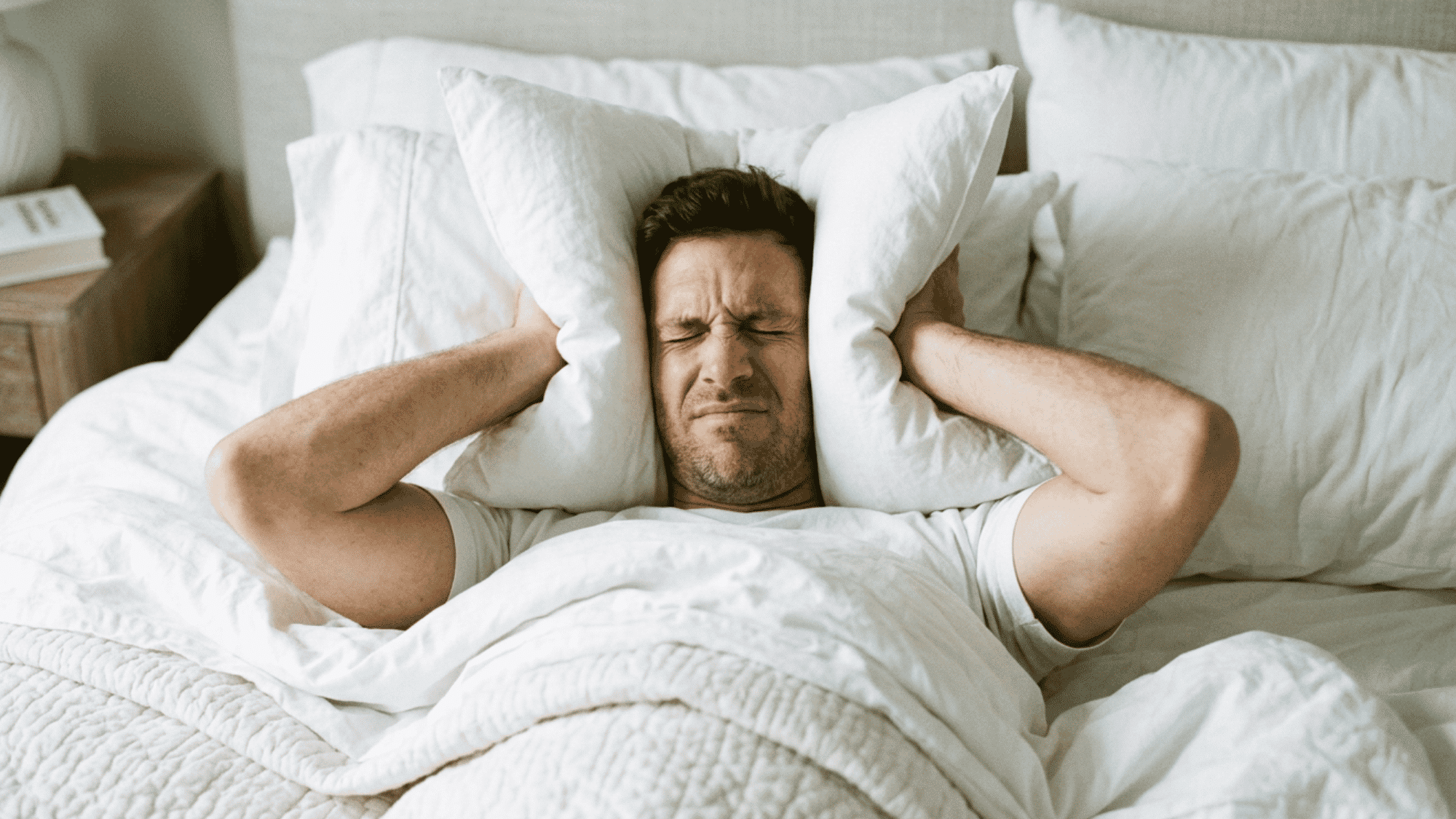 Person lying in bed covering ears with pillows while experiencing disturbed sleep.