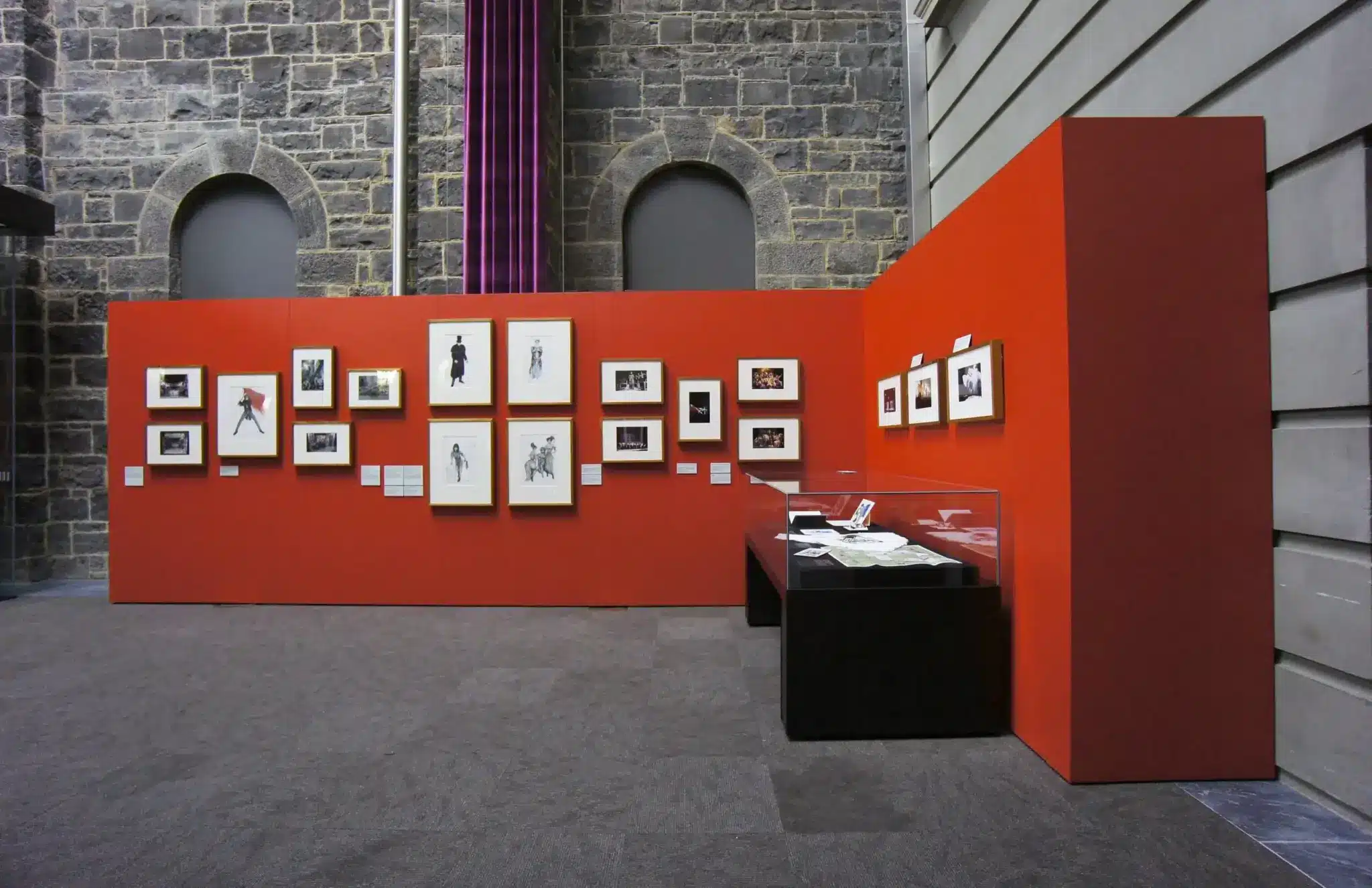 Art gallery exhibit with framed artwork on red walls and display case under soft lighting