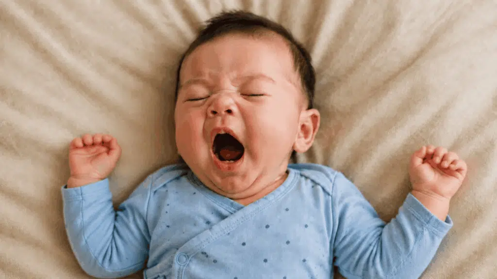 Yawning baby in a blue dotted outfit lying on soft beige bedding.