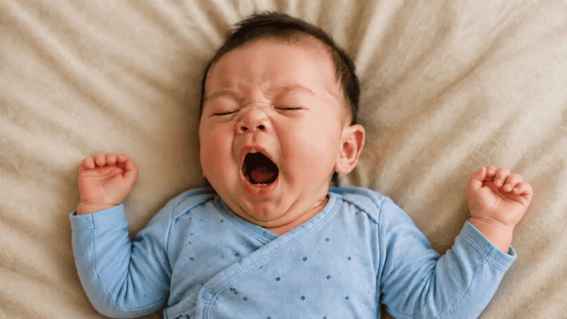 Yawning baby in a blue dotted outfit lying on soft beige bedding.