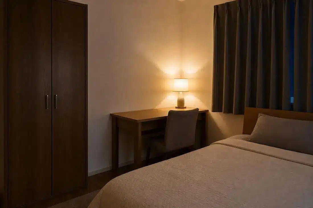 Cozy bedroom with wooden desk and lamp, soft lighting, beige bedding, and dark curtains