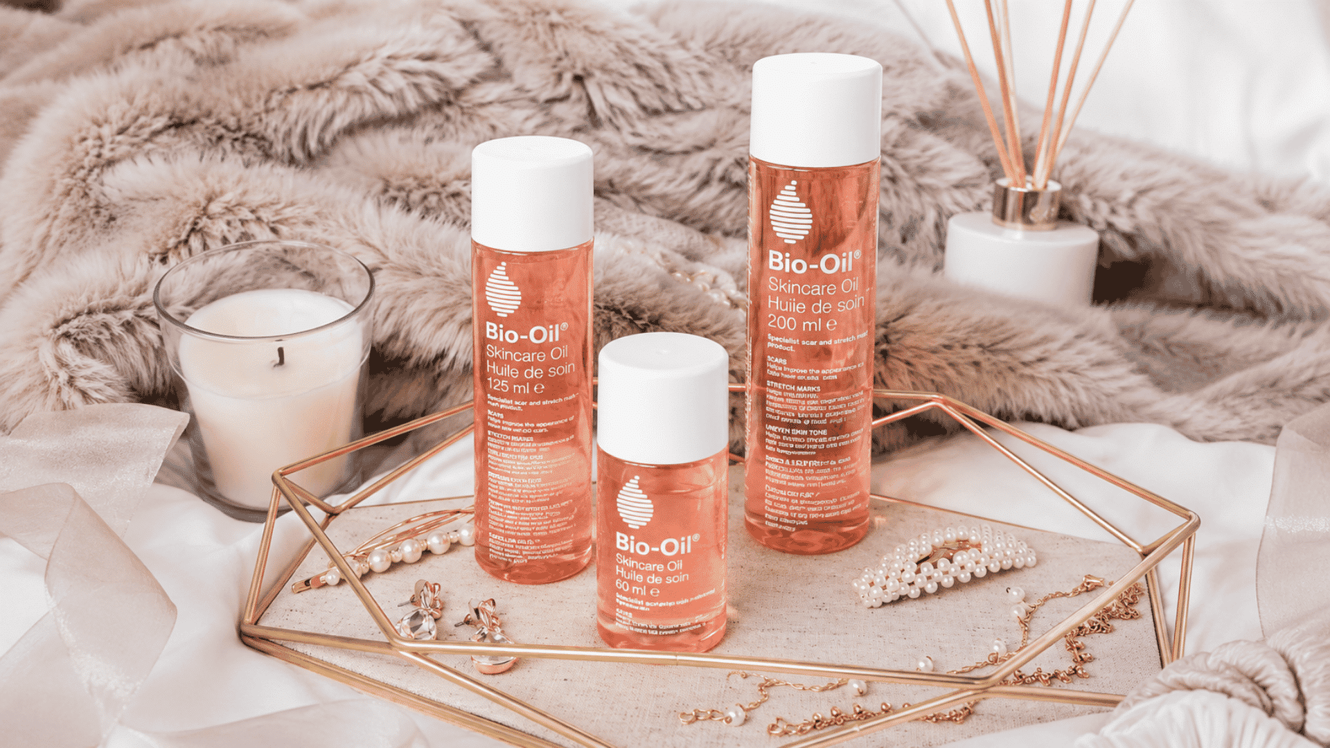 Three Bio-Oil skincare oil bottles arranged on a gold tray with pearls, a candle, and soft neutral décor.