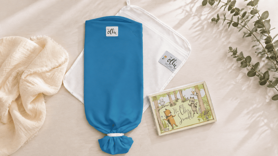 blue ollie swaddle displayed with its mesh pouch and packaging on a neutral background