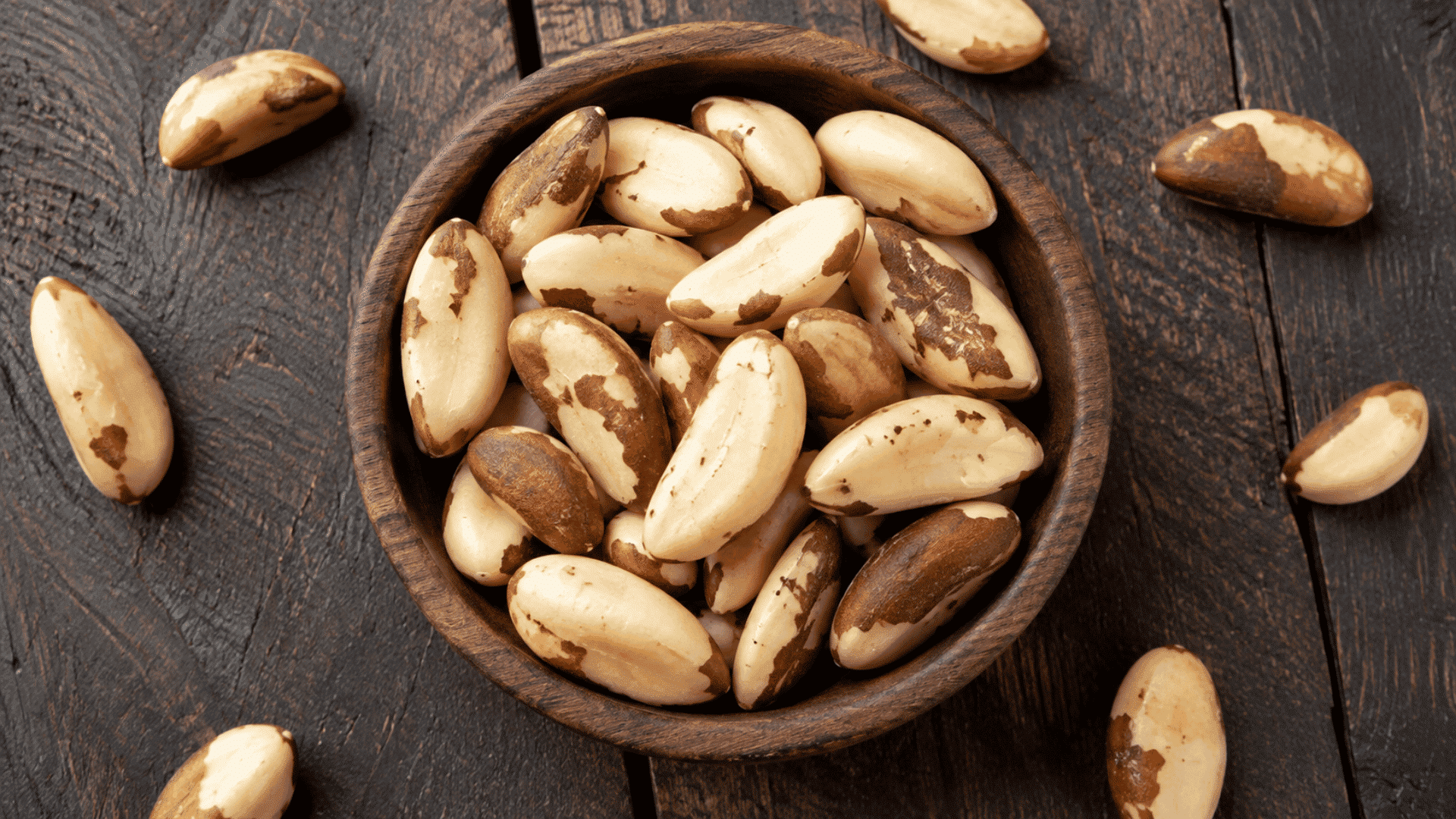 brazil nuts in a wooden bowl scattered across a dark rustic table