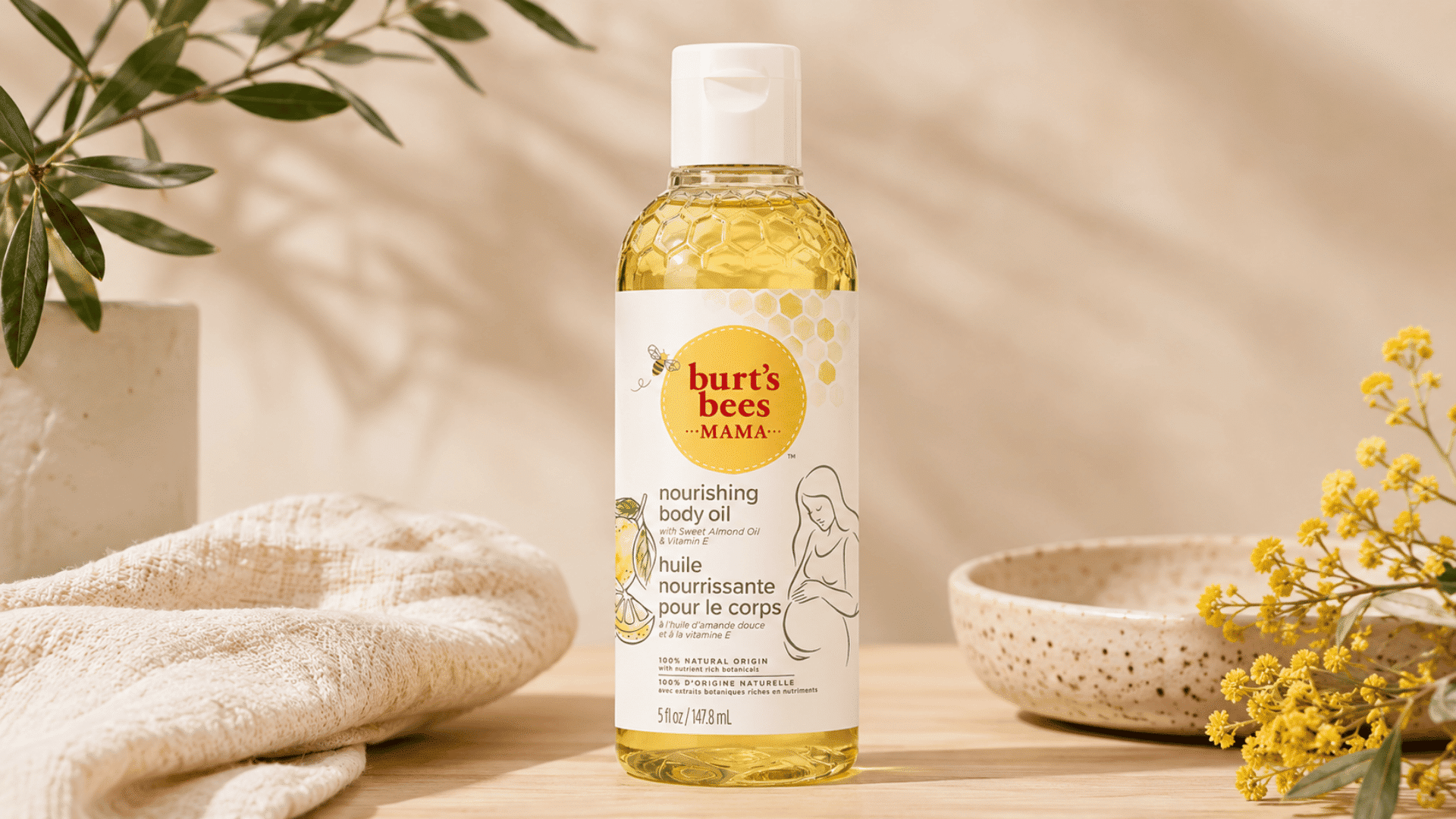 Burt’s Bees Mama Nourishing Body Oil bottle displayed with a cream towel, greenery, ceramic dish, and yellow flowers.