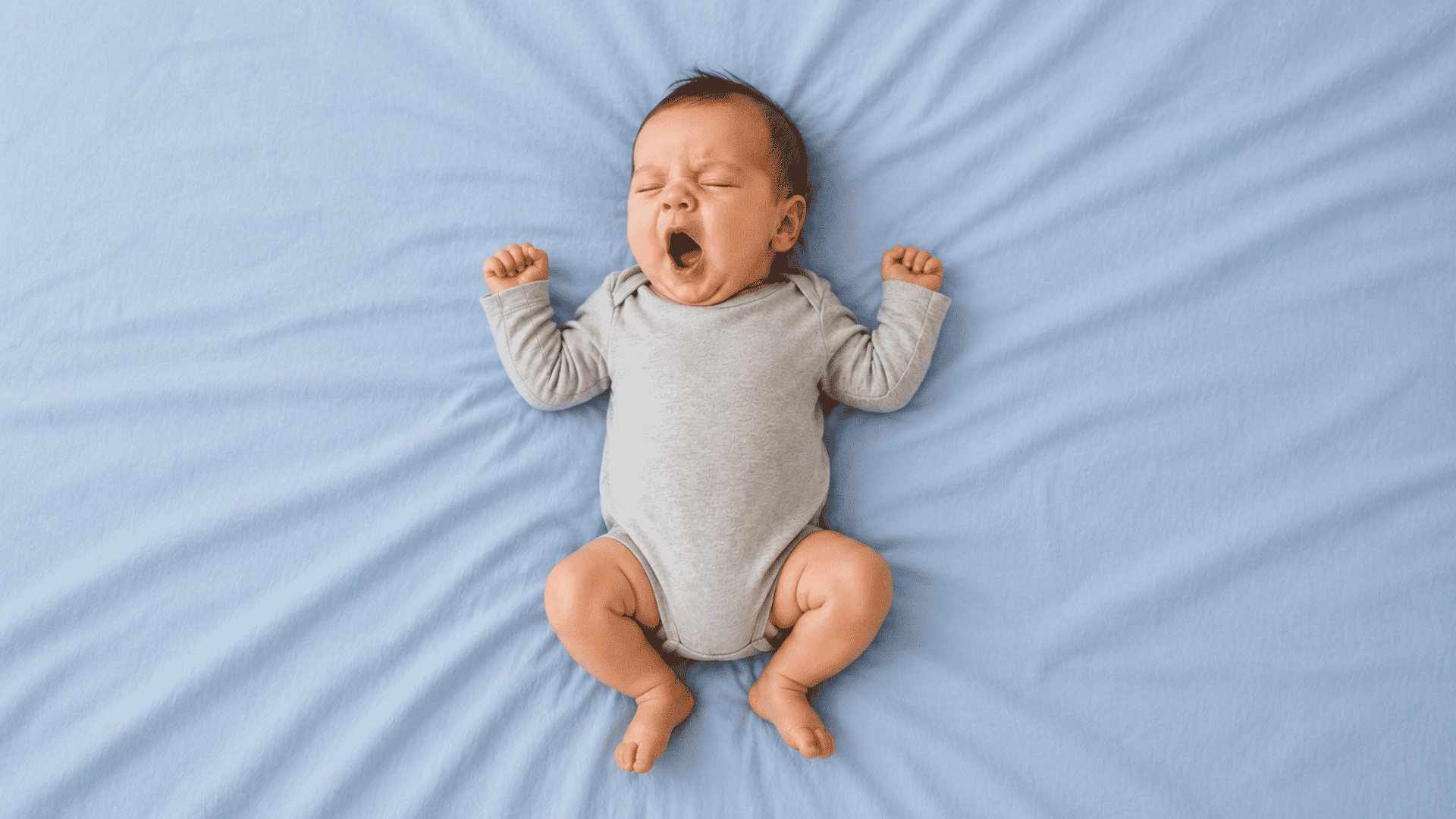 baby in a gray onesie yawning with arms stretched overhead on a light blue bed sheet