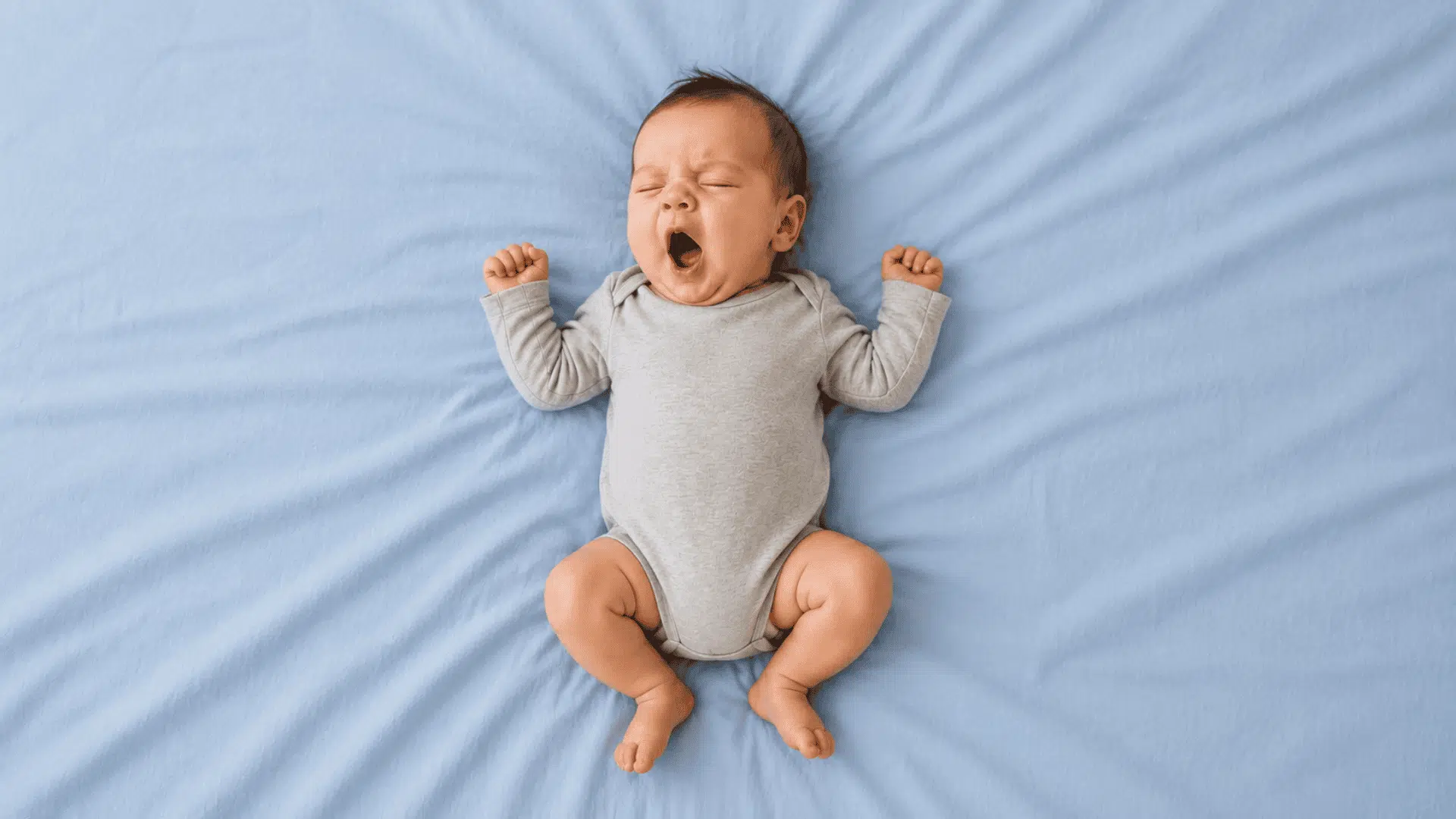 baby in a gray onesie yawning with arms stretched overhead on a light blue bed sheet