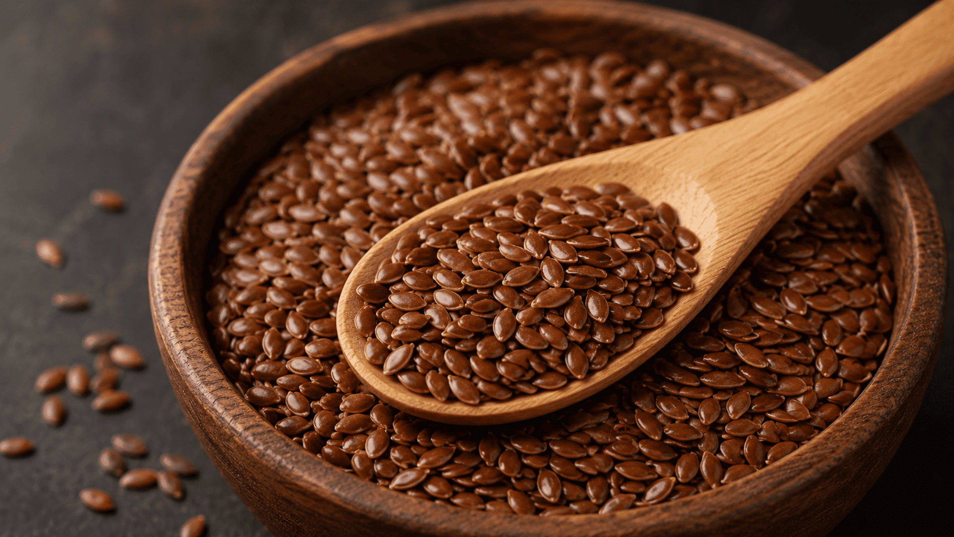flaxseeds in a wooden bowl with a filled wooden spoon on a dark surface