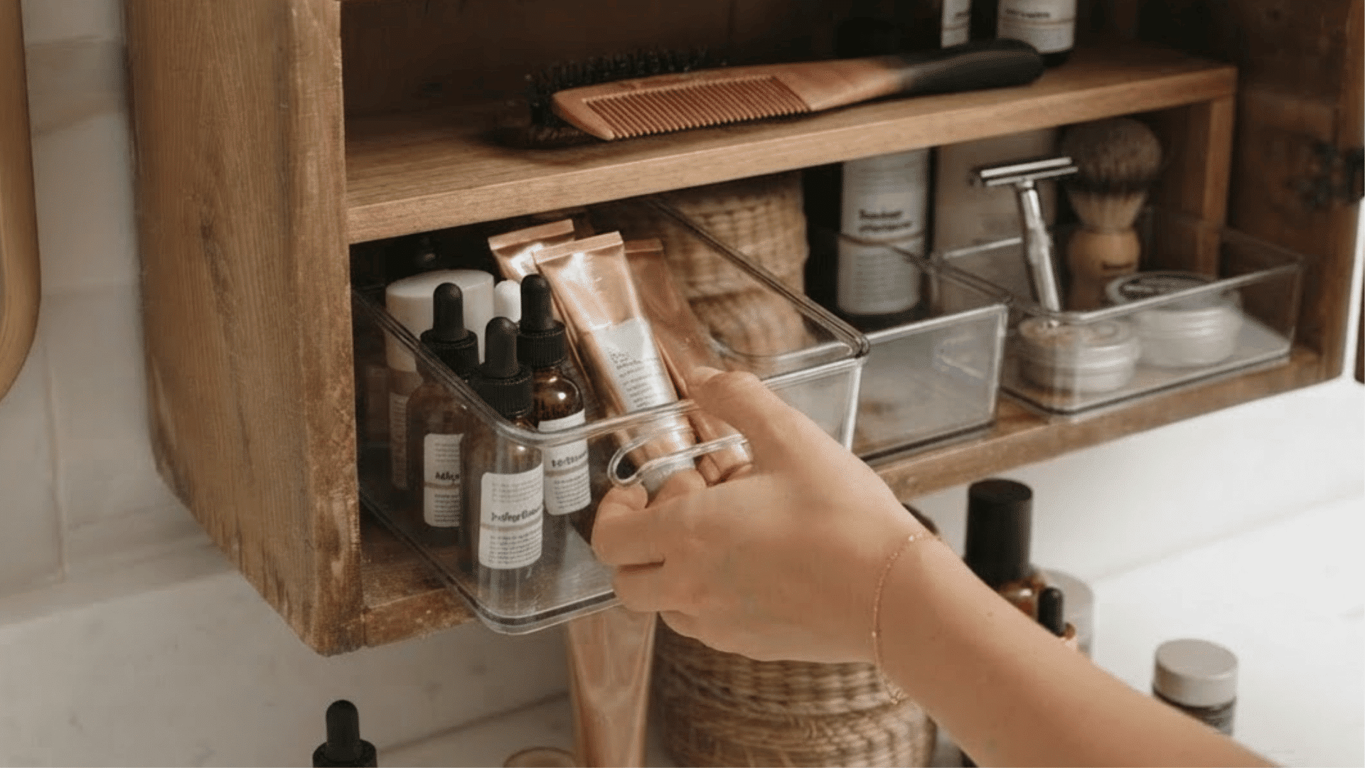 hand arranging skincare products in clear storage bins inside a wooden bathroom cabinet