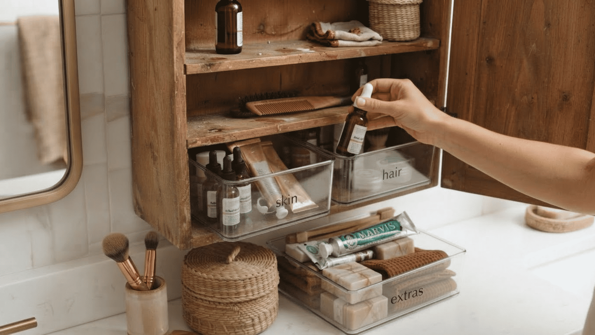 hand placing skincare bottle into labeled clear bins inside an organized bathroom cabinet
