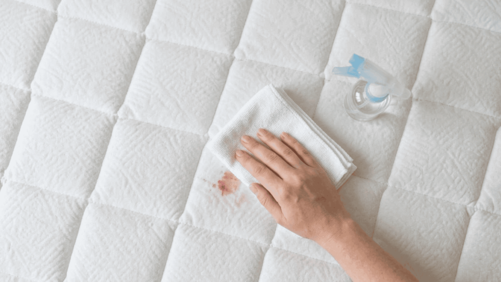 hand blotting a fresh blood stain on a white mattress with a cloth and spray bottle nearby