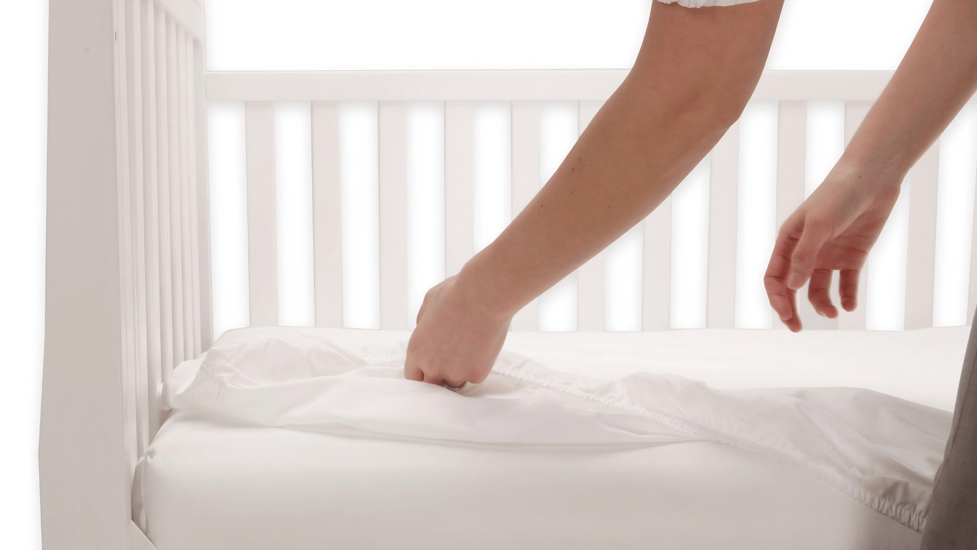 Caregiver smoothing a fitted sheet over a firm mattress in an empty white baby crib.