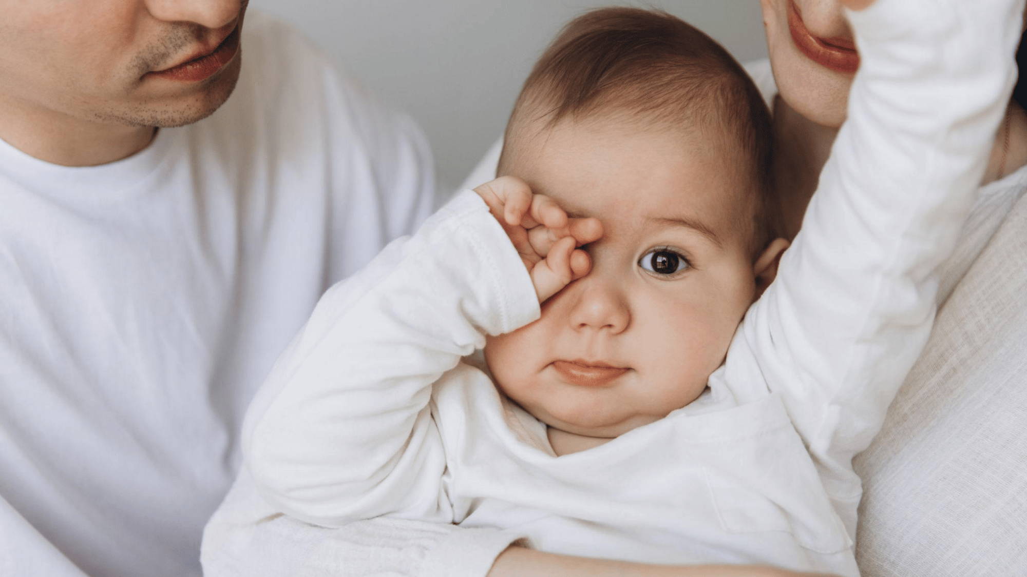 Tired baby rubbing one eye while being held between two parents.